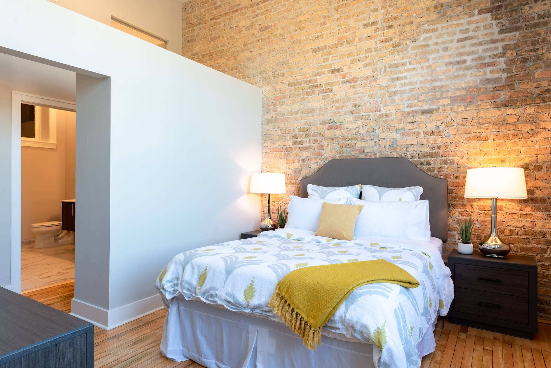 Main bedroom with wood flooring, large window, high ceiling, exposed brick wall, and doorway to walk-in closet and adjoining bathroom.
