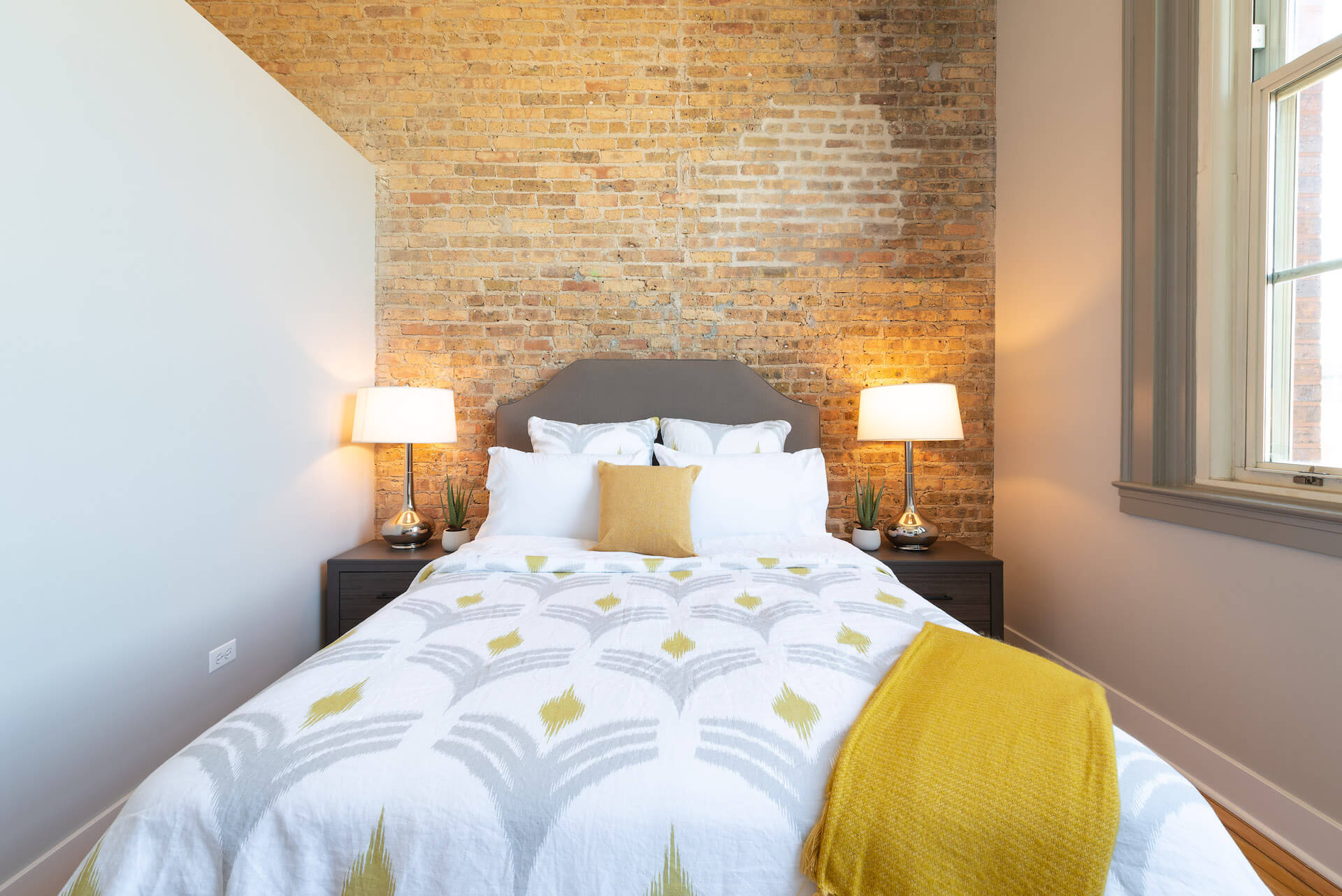 Main bedroom with wood flooring, large window, high ceiling, and exposed brick wall.