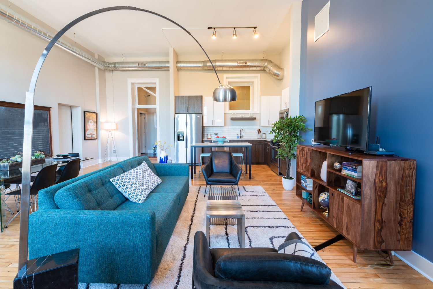 Living room with blue accent wall, high ceiling, wood style flooring, completely open to kitchen and dining space.